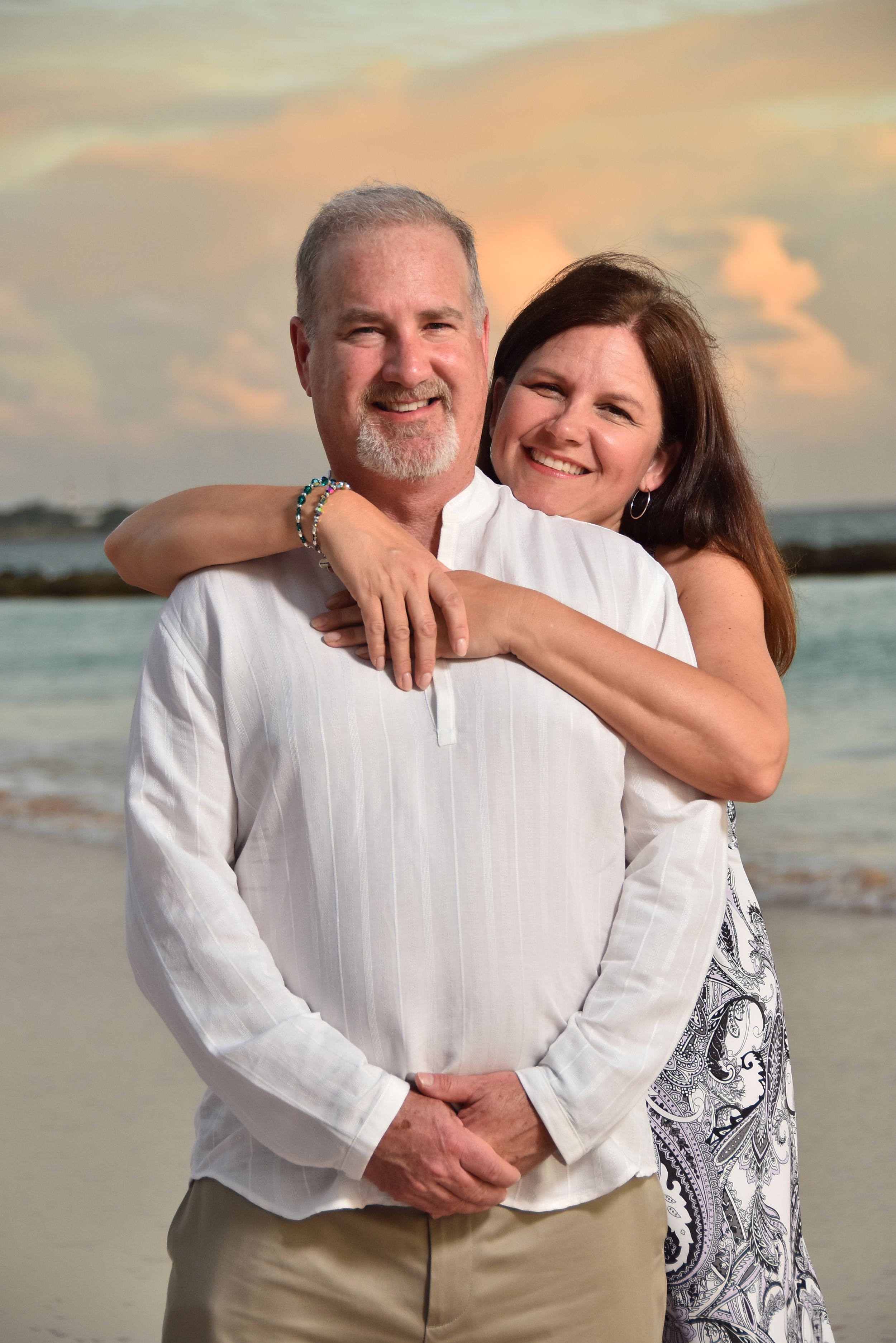 Couple in white clothing embraces on beach at sunset with ocean and warm sky in background.