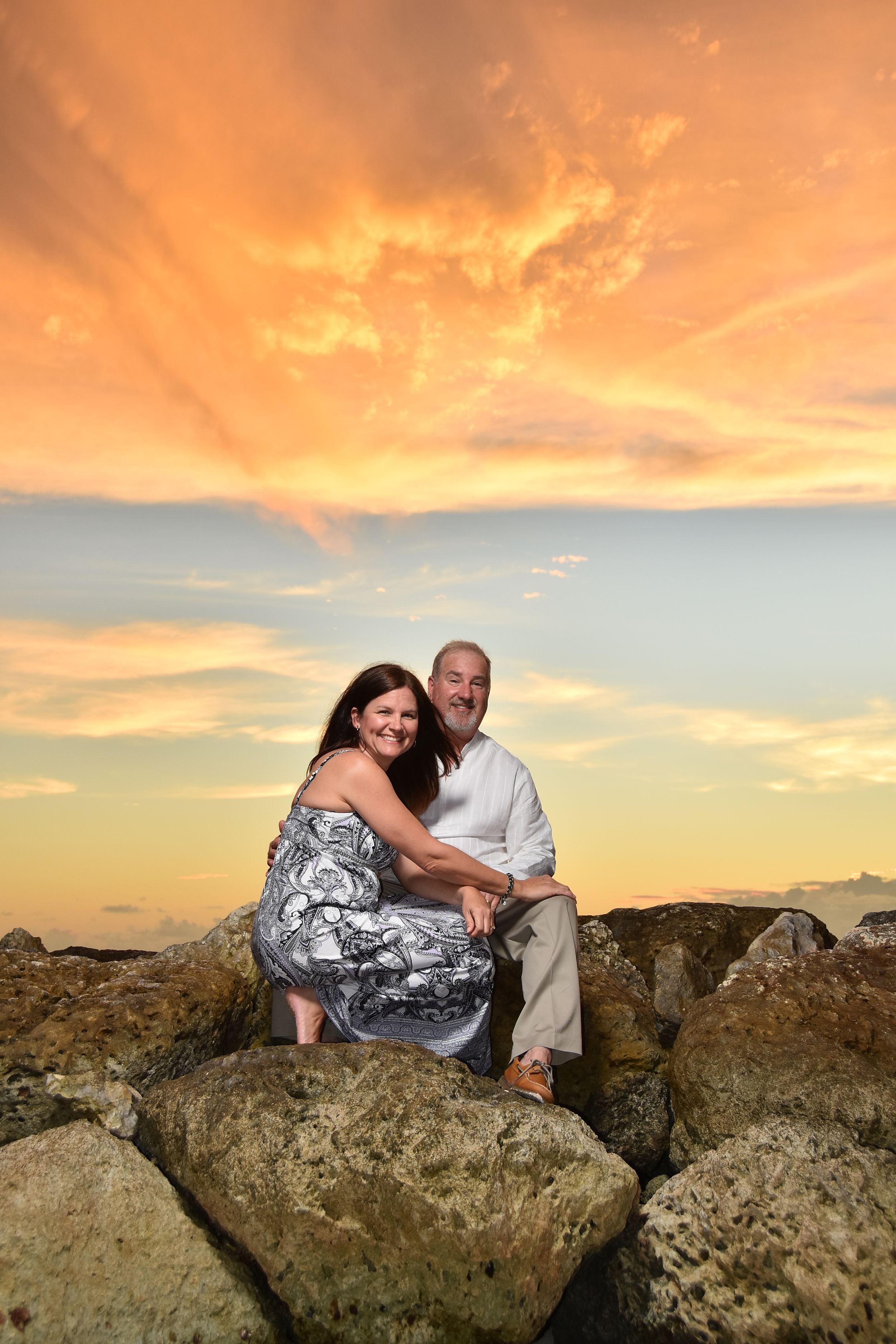 Couple sits together on coastal rocks during a dramatic orange and pink sunset.