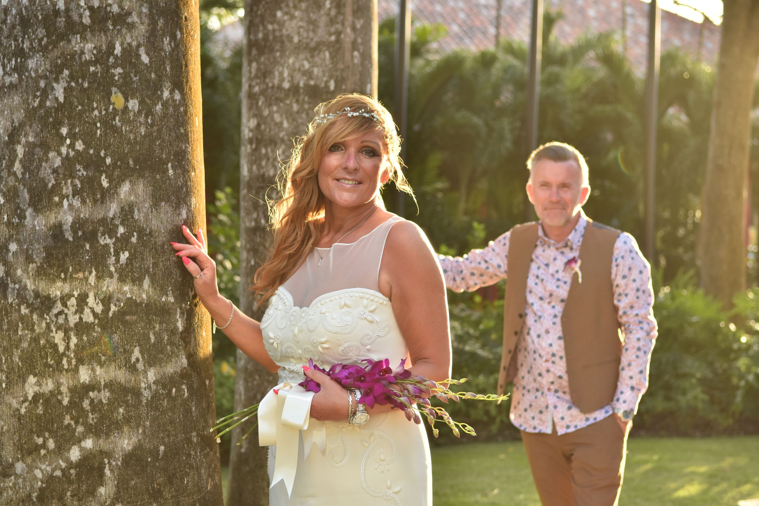 Wedding couple stands by large tree trunk with purple bouquet in warm evening light.