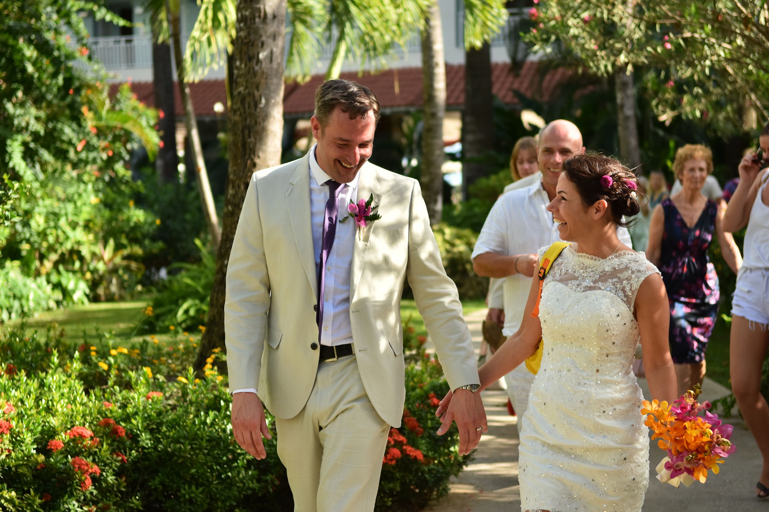 A wedding couple walks together on a garden path surrounded by lush greenery and tropical flowers.