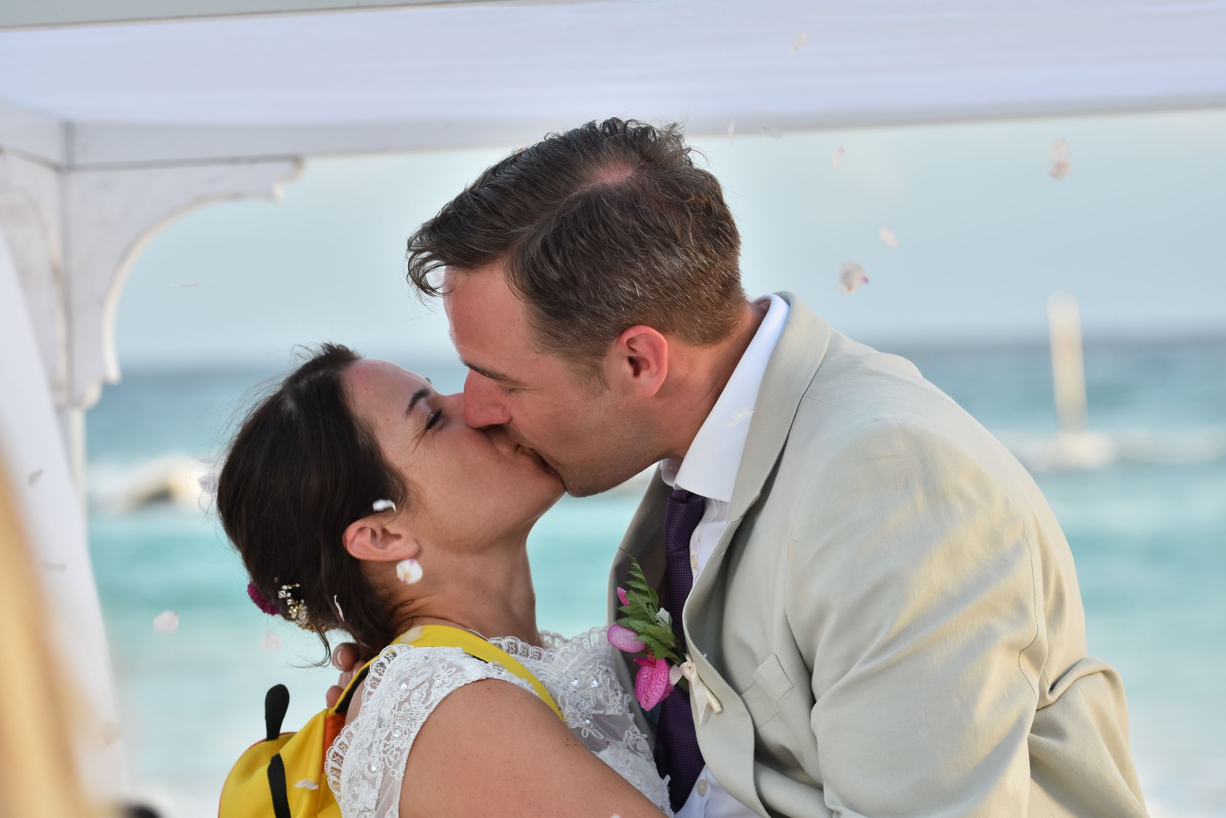 A couple shares a romantic kiss under a beach canopy with turquoise ocean waters in the background.