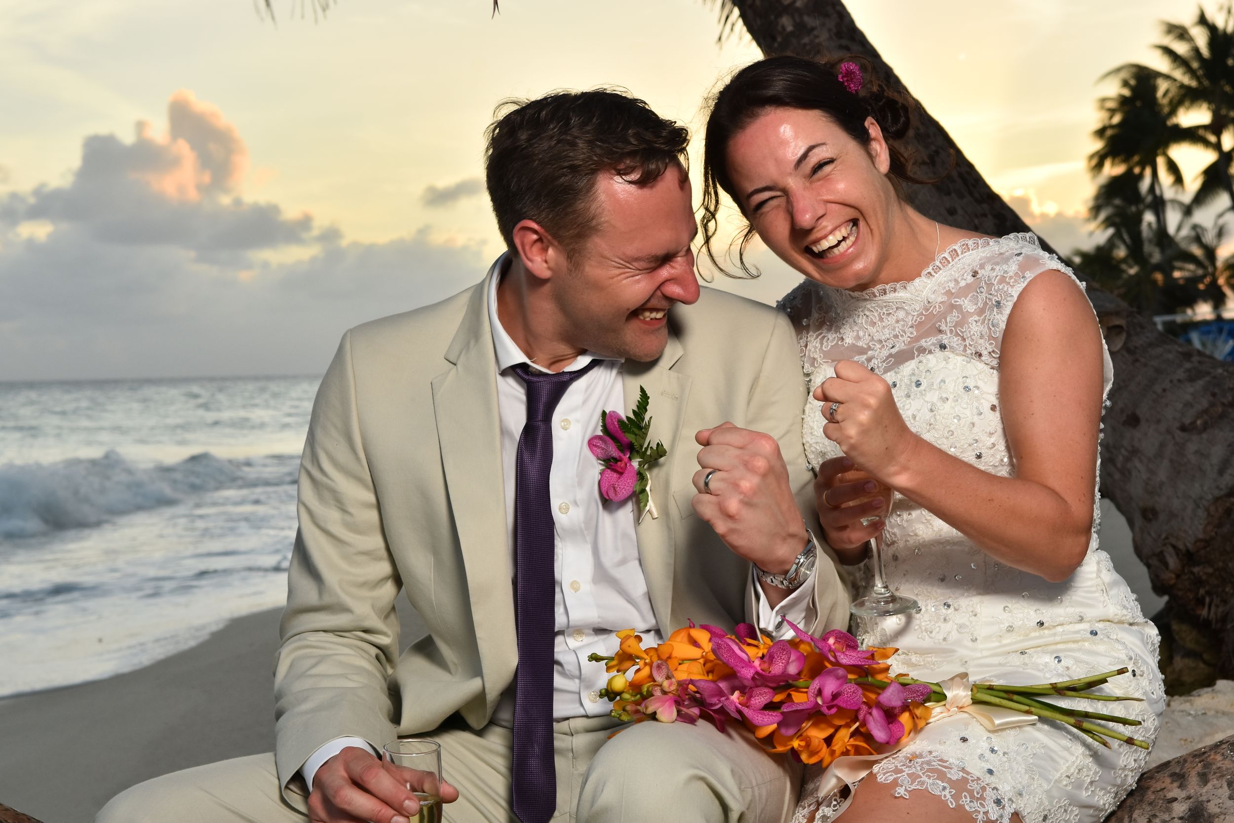 A couple shares a joyful moment together on a beach at sunset with colorful tropical flowers.