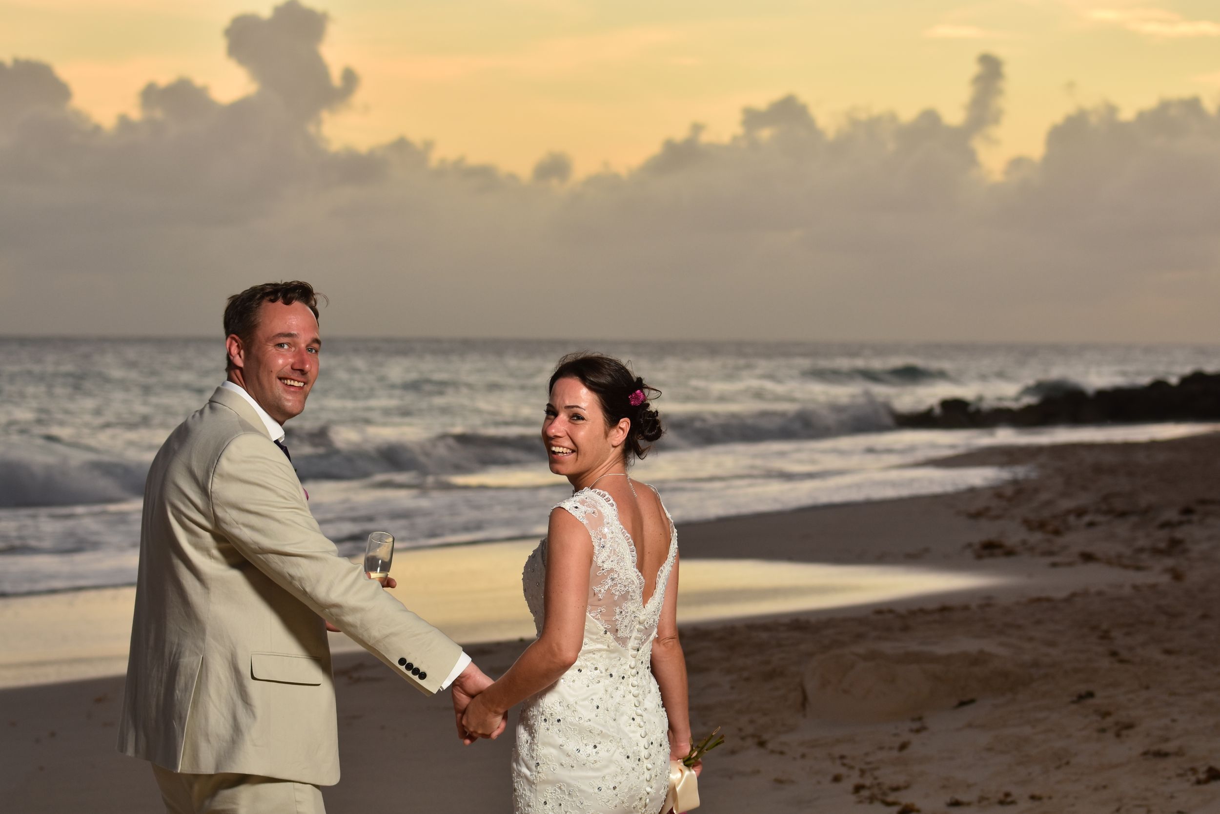 A couple shares a joyful moment at sunset on a beach, with a cloudy sky and ocean waves in the background.