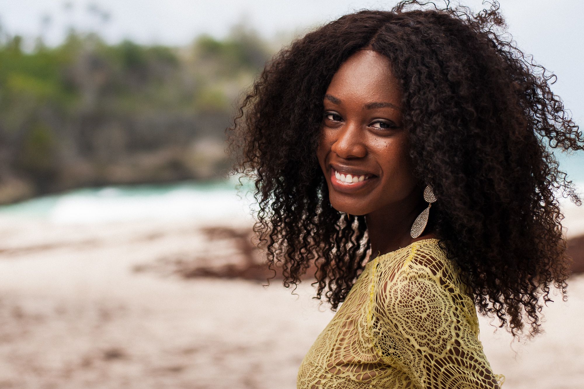 A woman with natural curly hair wearing a yellow knit sweater smiles while posing on a beach.