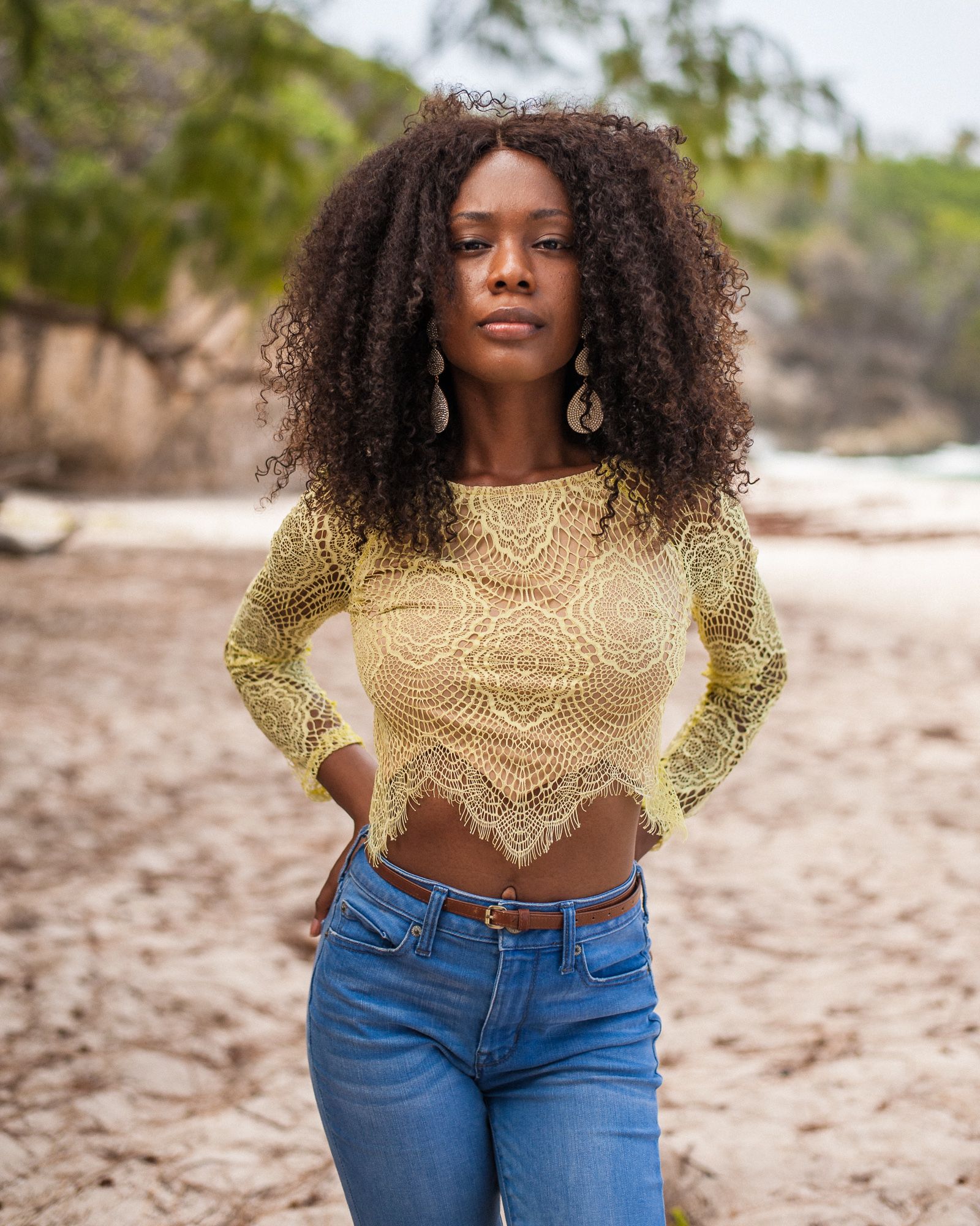 A person in a yellow lace crop top and blue jeans poses on a beach with natural curly hair and silver earrings.