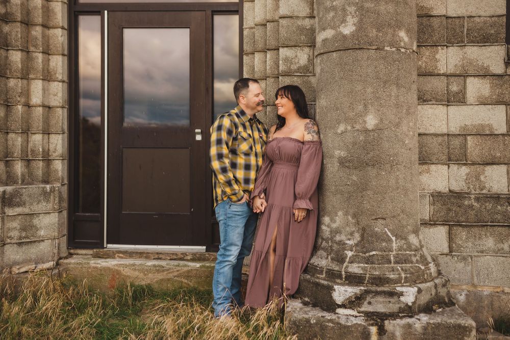 A couple in casual attire stands together beside stone pillars of a rustic building with a dark wooden door.