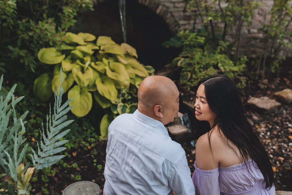 A romantic couple in casual attire share an intimate moment together in a garden with lush plants and stone walls.