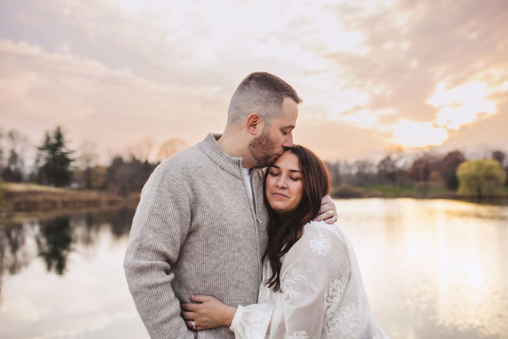 Couple embracing by tranquil lake at sunset with reflection on water and cloudy sky in background.
