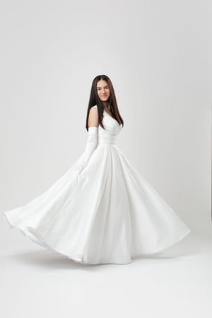 Elegant bride in flowing white ball gown and gloves, twirling gracefully against a soft white studio background.