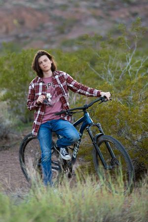 Teen boy with long hair in plaid shirt and jeans posing with mountain bike on desert trail.