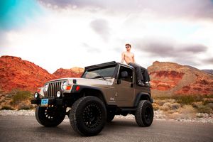 Shirtless man standing through open roof of tan Jeep Wrangler on desert road with red rock formations.