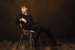 A person in all black attire sits casually on a chair against a dark brown backdrop on wooden flooring.