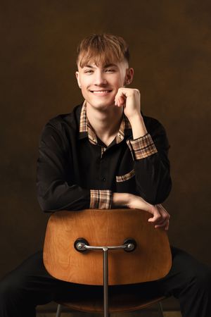 A person in a black sweater with plaid details sits casually posed on a brown wooden chair against a dark background.