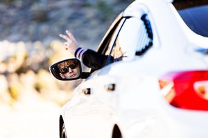 A reflection in white car's side mirror shows sunglasses against a blurred mountain background.