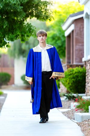 A graduate poses in a blue graduation gown while walking on a sidewalk in front of brick buildings and greenery.