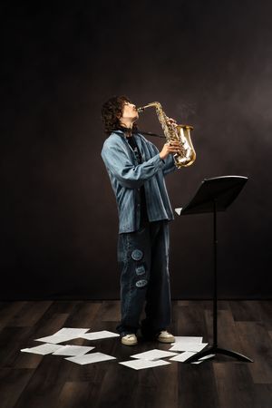 Young musician in denim plays saxophone with sheet music scattered on wooden floor during moody performance.