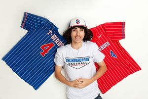 Baseball player wearing white shirt poses between blue and red pinstriped jerseys displayed on wall.