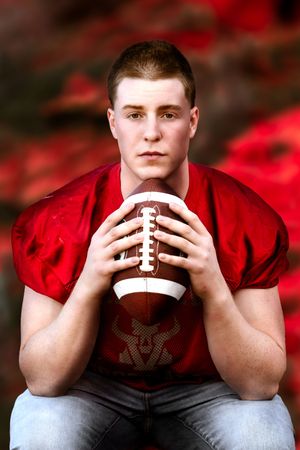 Young male football player in red jersey holding a football, posing seriously against a red autumn background.