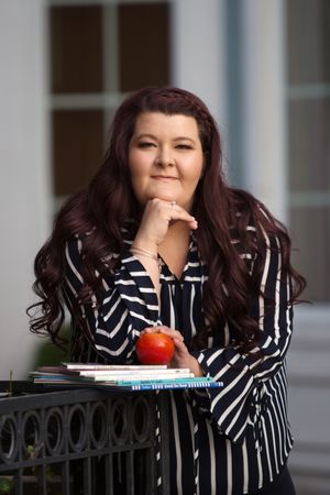 Woman with dark red hair in striped shirt poses with books and apple, smiling confidently outdoors.