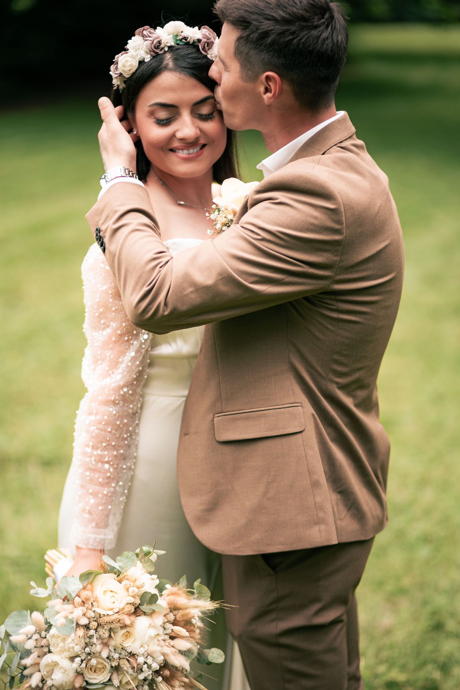 Groom in tan suit kissing bride wearing floral crown and holding bouquet outdoors at wedding ceremony.