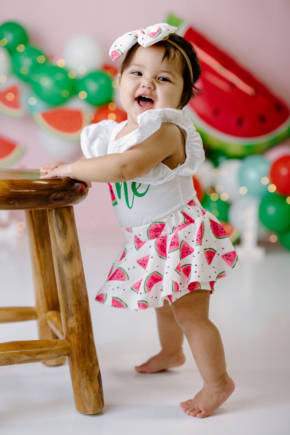 Baby in watermelon print dress holding wooden stool with fruit-themed party decorations in background.