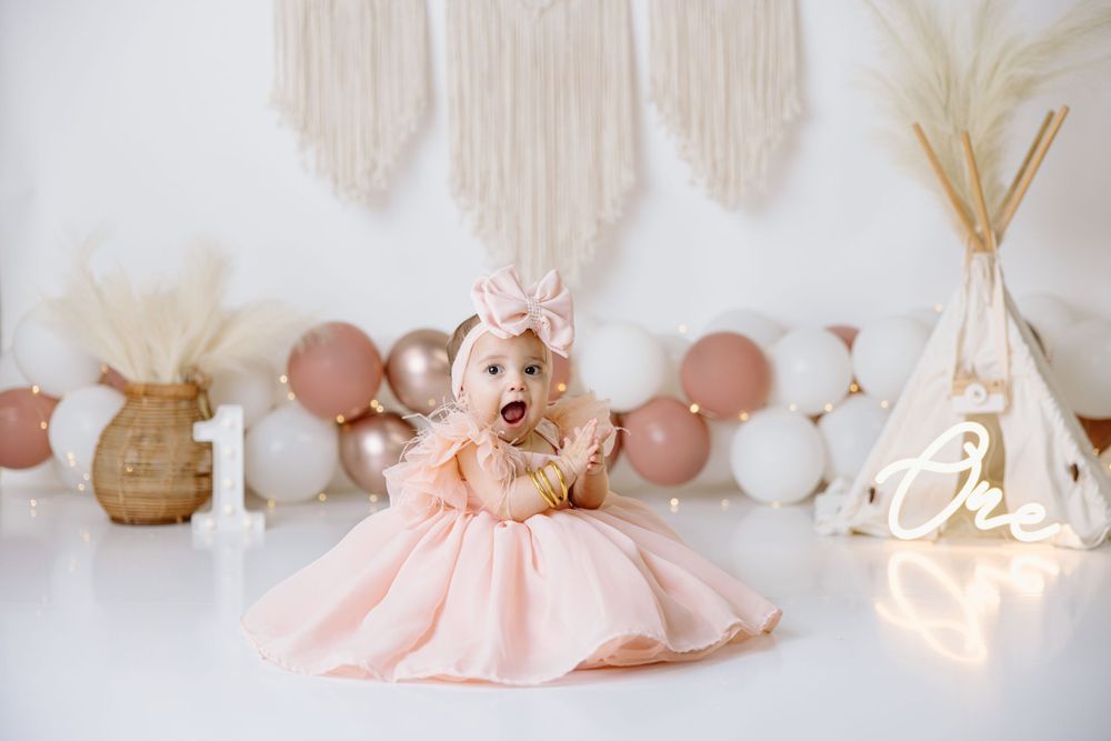 Baby girl in pink dress sitting among boho decorations with balloons and macrame backdrop.