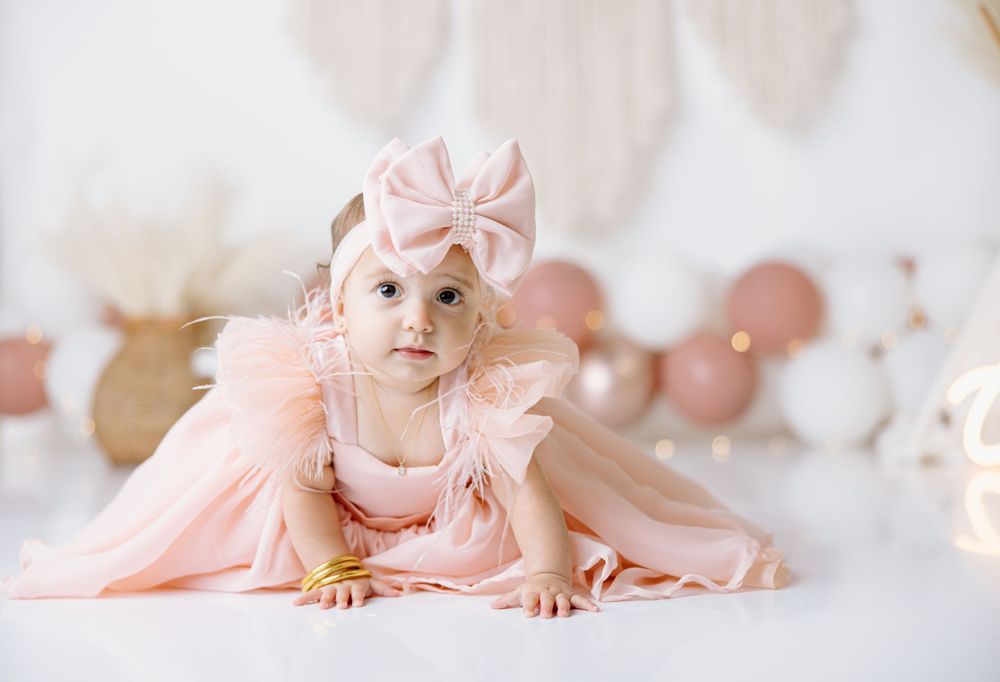 A baby wearing a pink tulle dress and bow headband poses on a light backdrop with pink balloons.