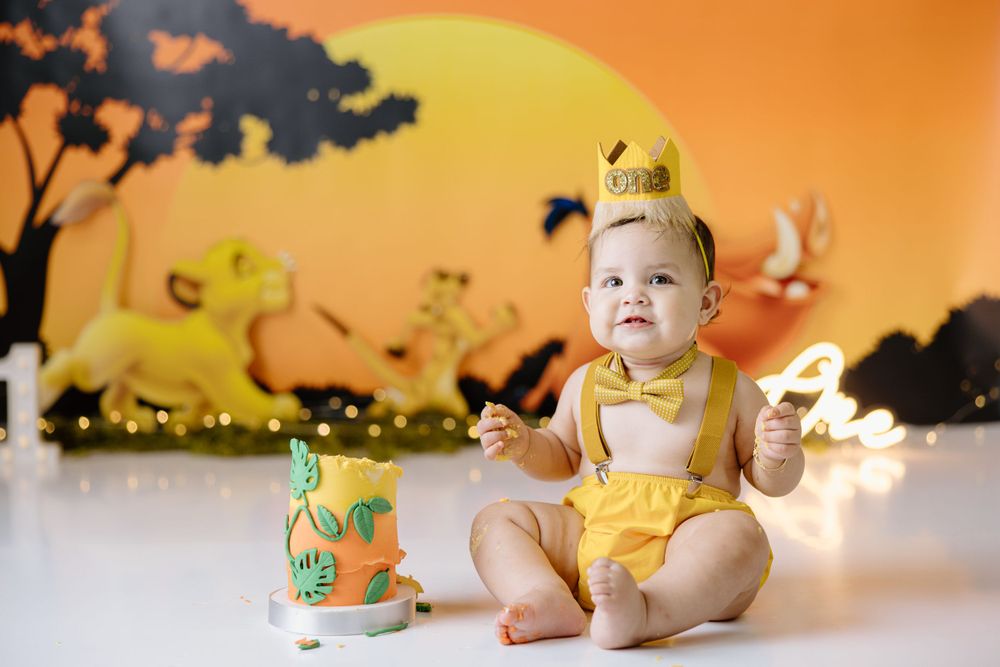 Baby in yellow outfit and crown sitting next to decorative cake against sunset themed backdrop.