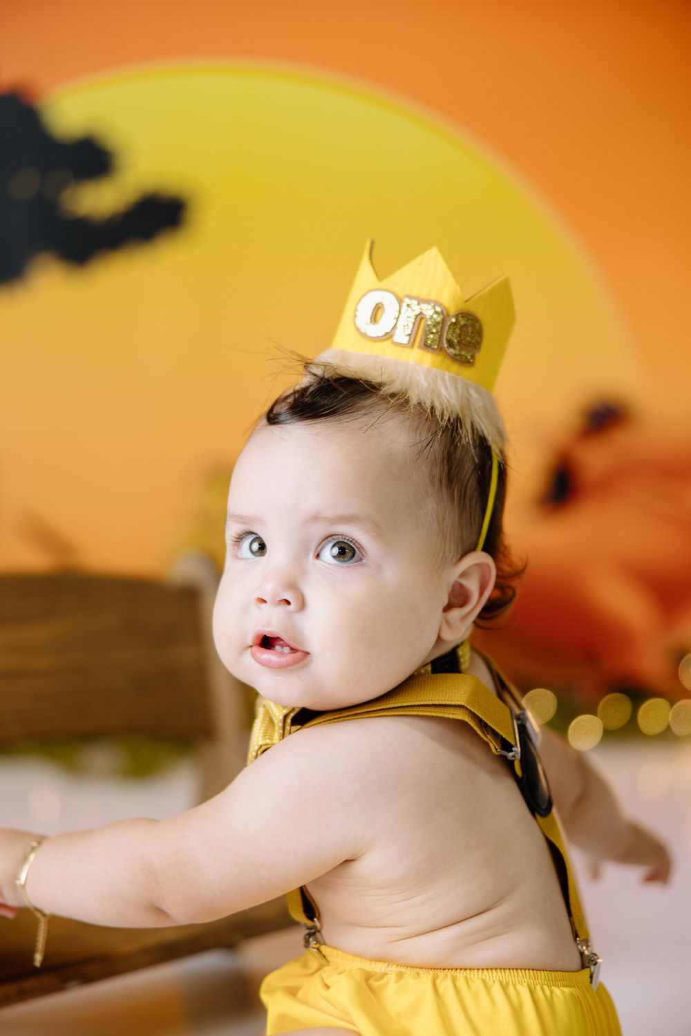 Baby wearing gold crown and yellow outfit against orange sunset backdrop smiling during photo shoot.
