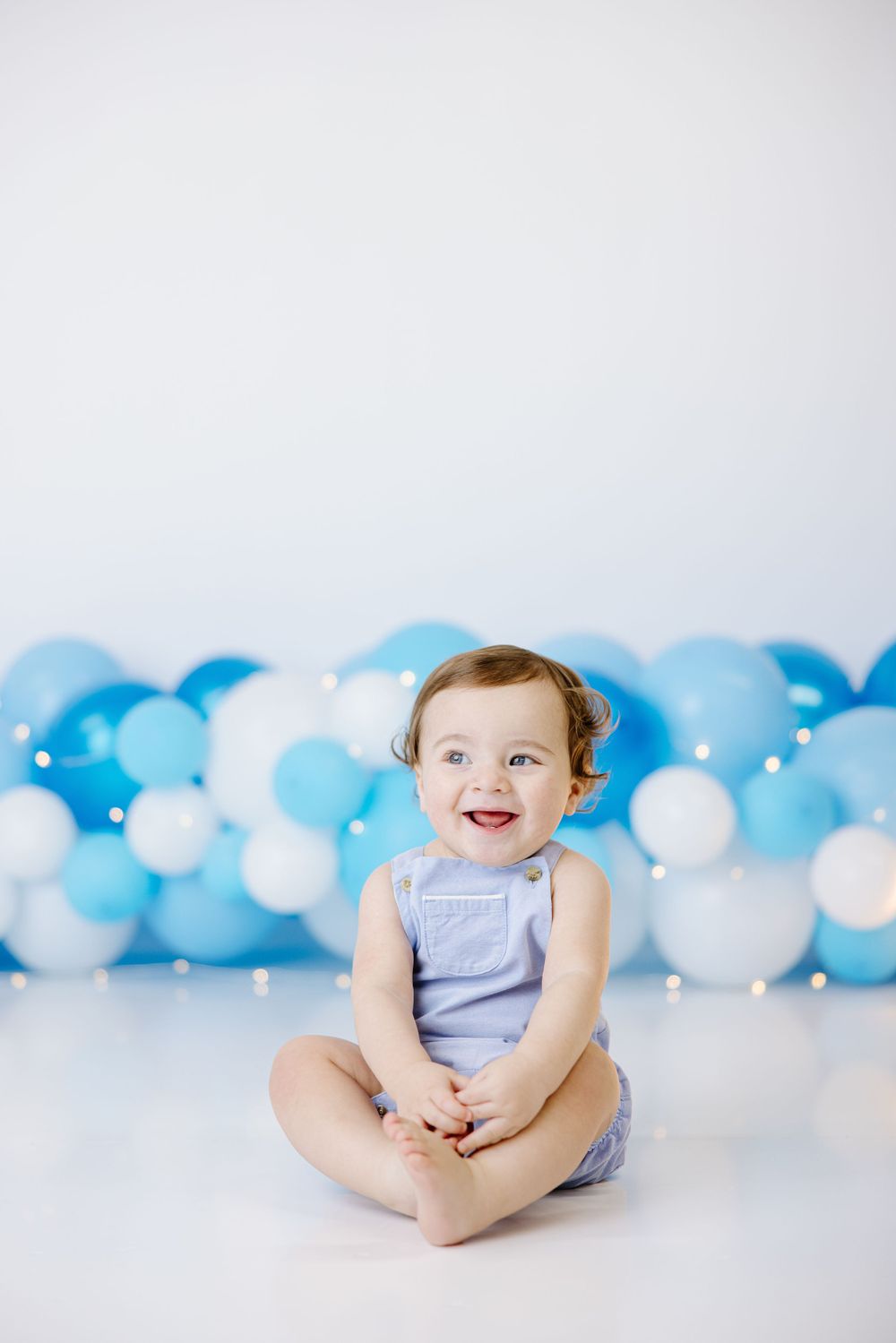 A baby in blue romper sits smiling against a backdrop of blue and white balloons.