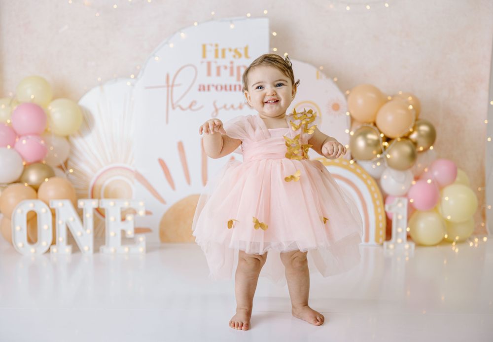 Baby in pink tulle dress smiles at first birthday photoshoot with balloon decorations.