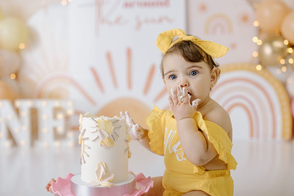 Baby in yellow outfit tastes birthday cake during celebratory photoshoot.
