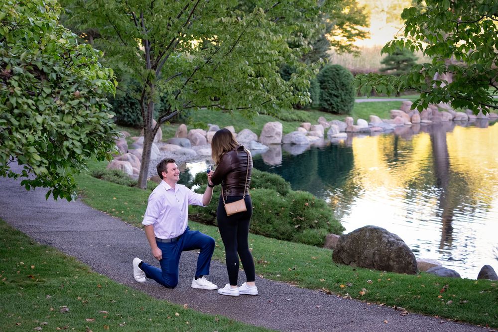 Man proposing to woman on one knee beside scenic garden pond.