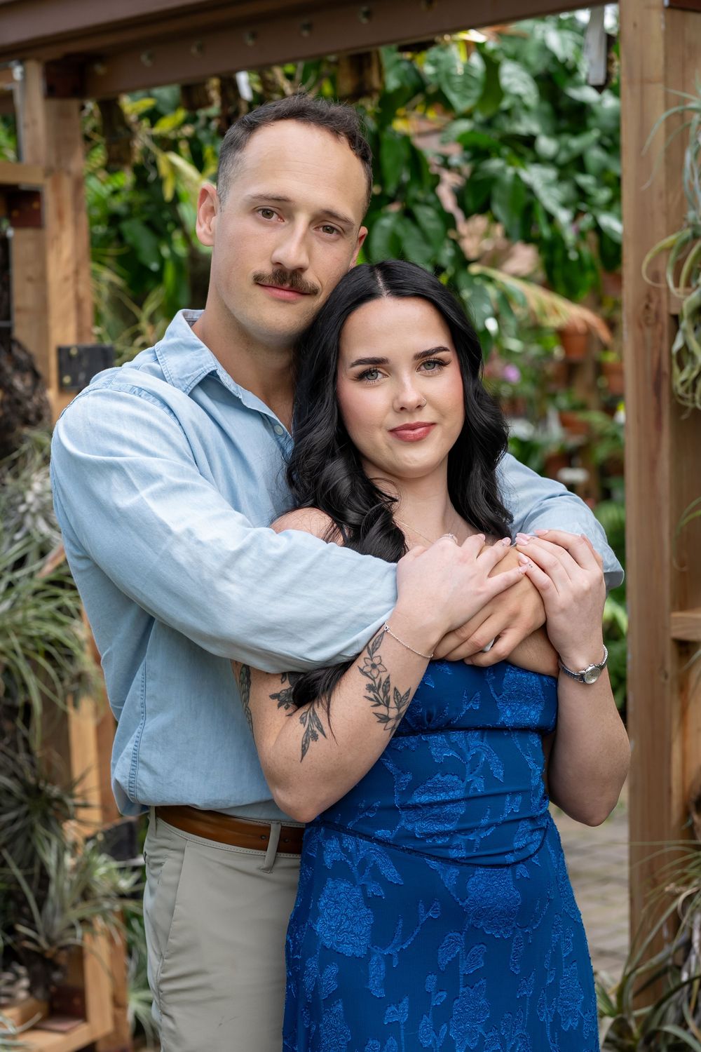 Happy couple embracing in lush greenhouse, man in blue shirt hugging woman in blue floral dress.