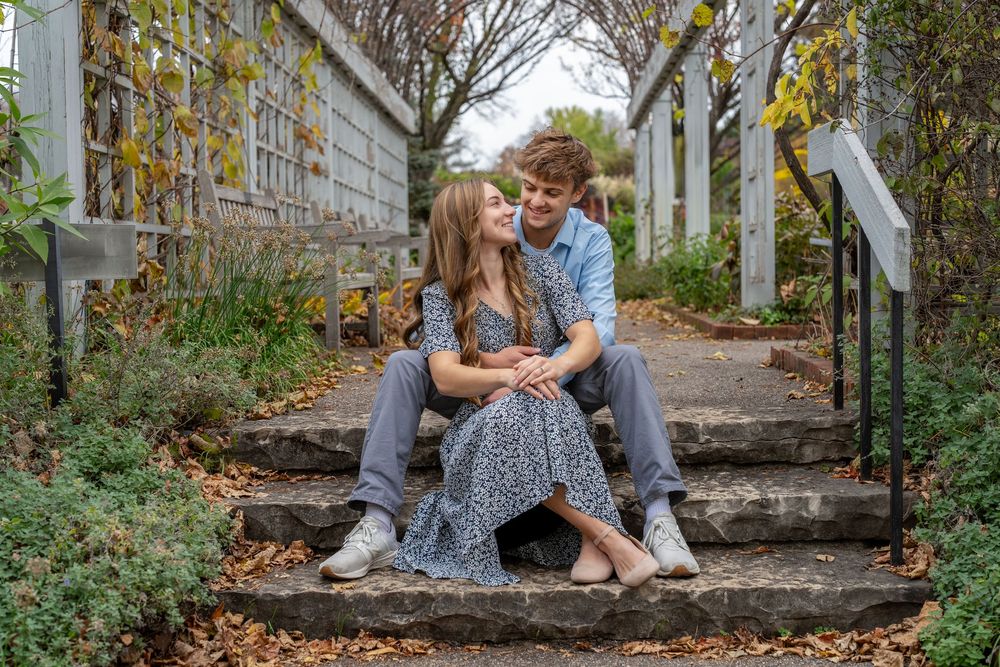 Smiling couple sits close together on rustic stone steps surrounded by greenery and autumn leaves in a romantic outdoor engagement photo.