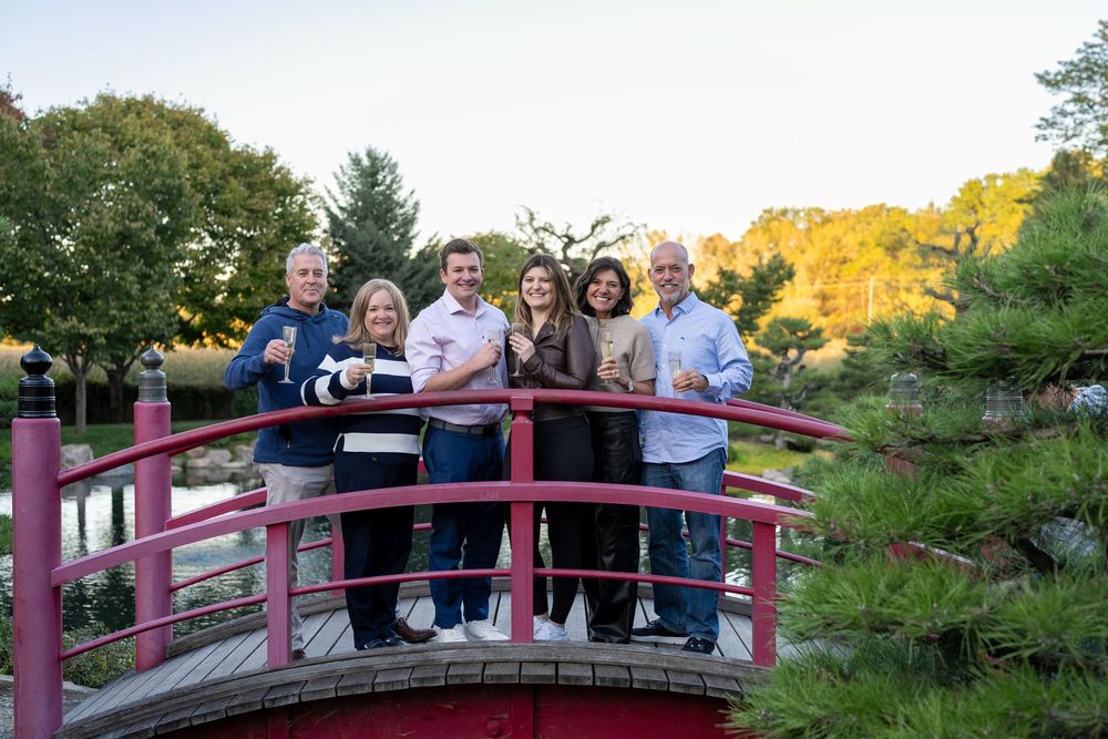 Group of six smiling people posing on a red arched bridge in a Japanese garden with lush green trees.