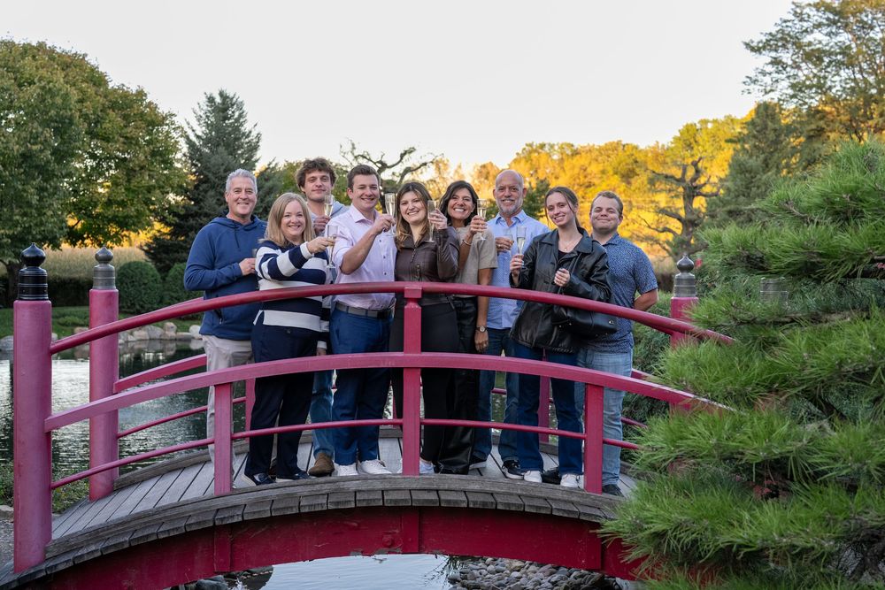 Group of nine people smiling on a red arched bridge surrounded by lush green trees in a Japanese garden.