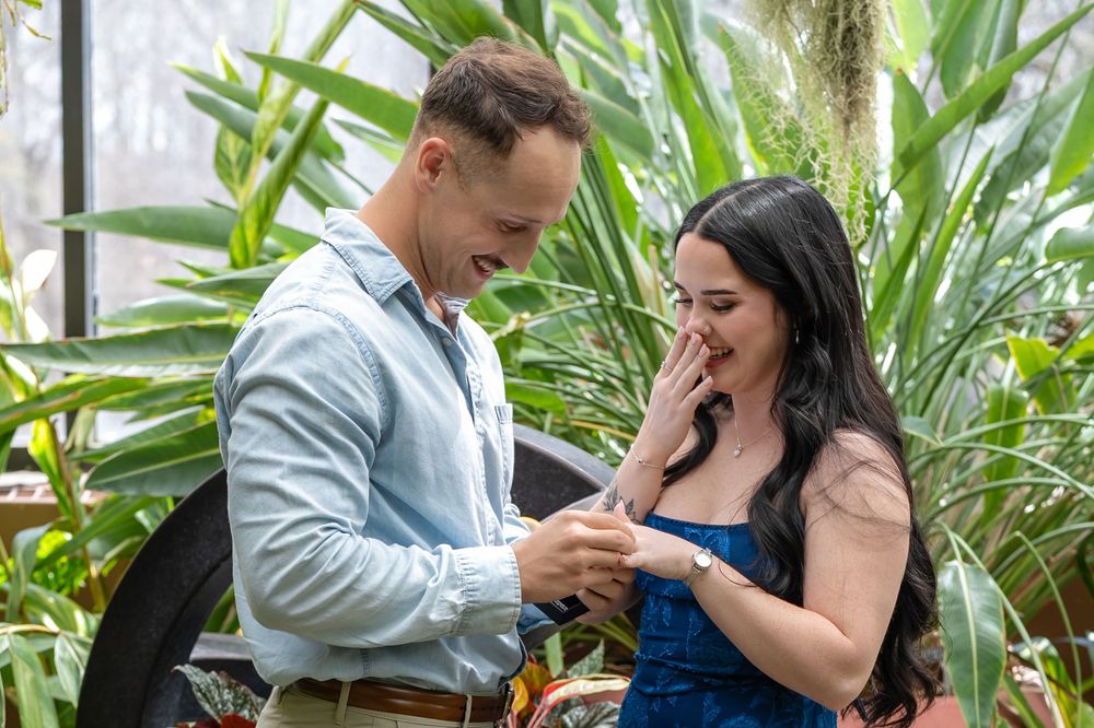 Joyful marriage proposal moment as man places ring on surprised womans finger in greenhouse.
