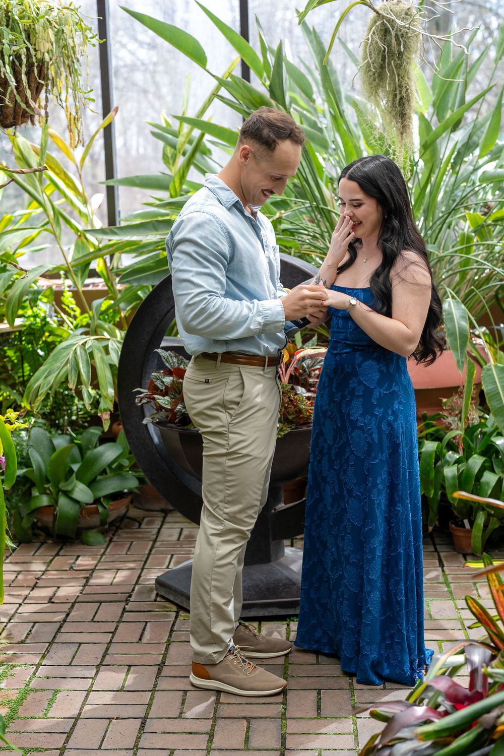 Couple sharing a tender engagement moment in greenhouse, man placing ring on womans finger.