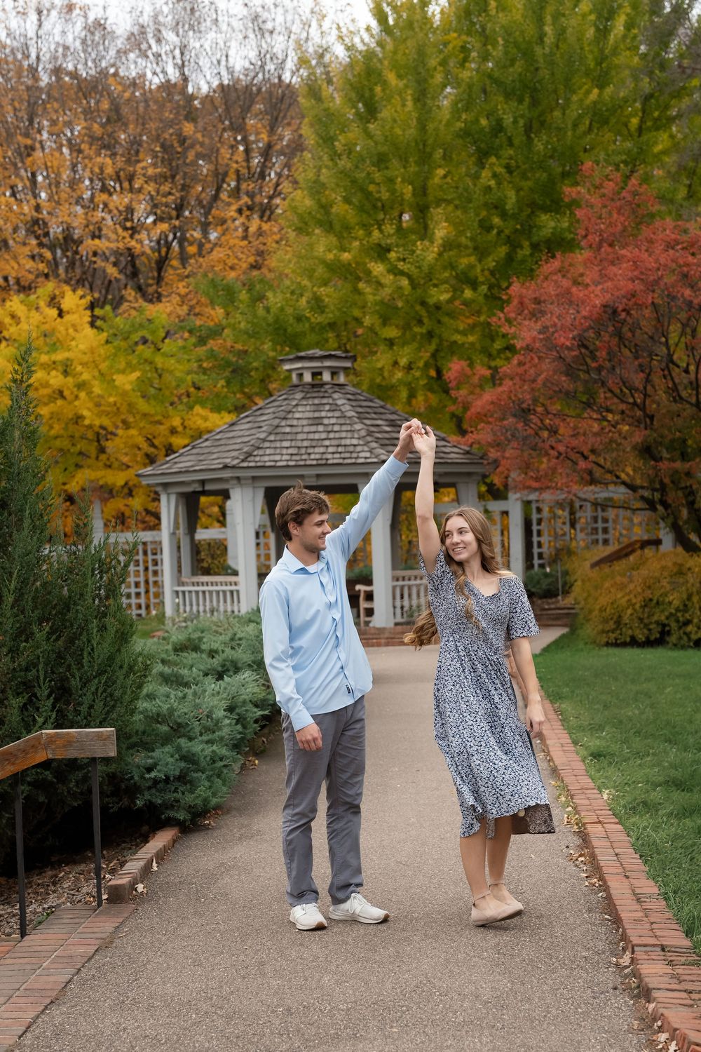 Young couple dancing hand in hand on a garden path near a wooden gazebo, surrounded by colorful autumn trees.