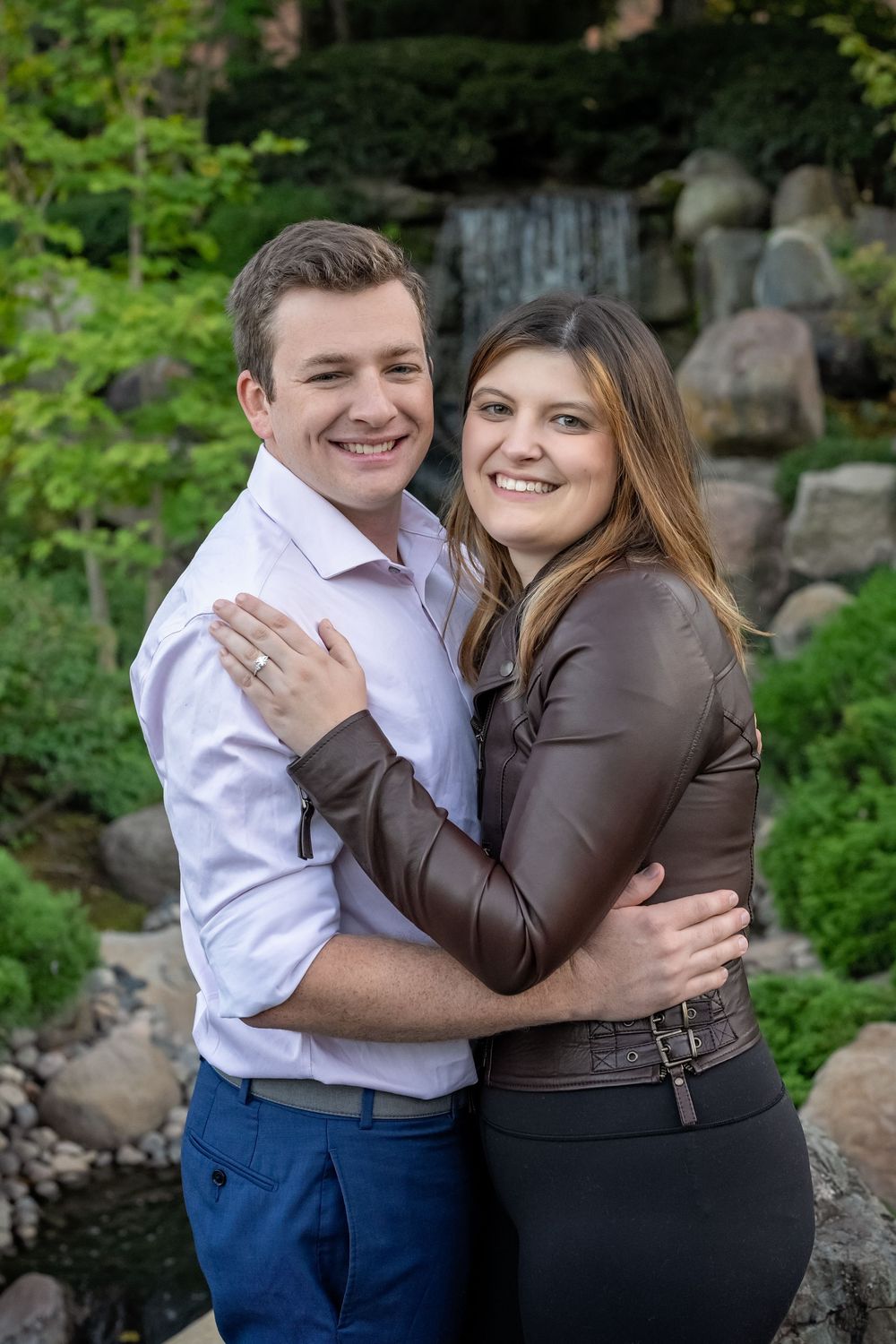 Smiling engaged couple hugging near waterfall and rocks in lush garden.
