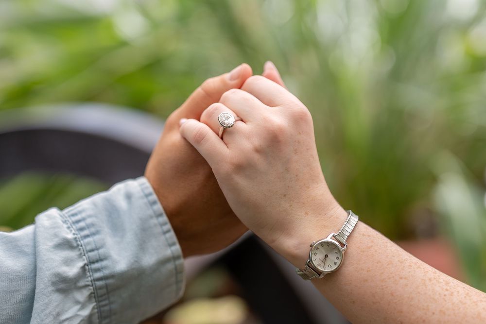 Close-up of hands showing engagement ring with diamond, one hand wearing a silver watch.