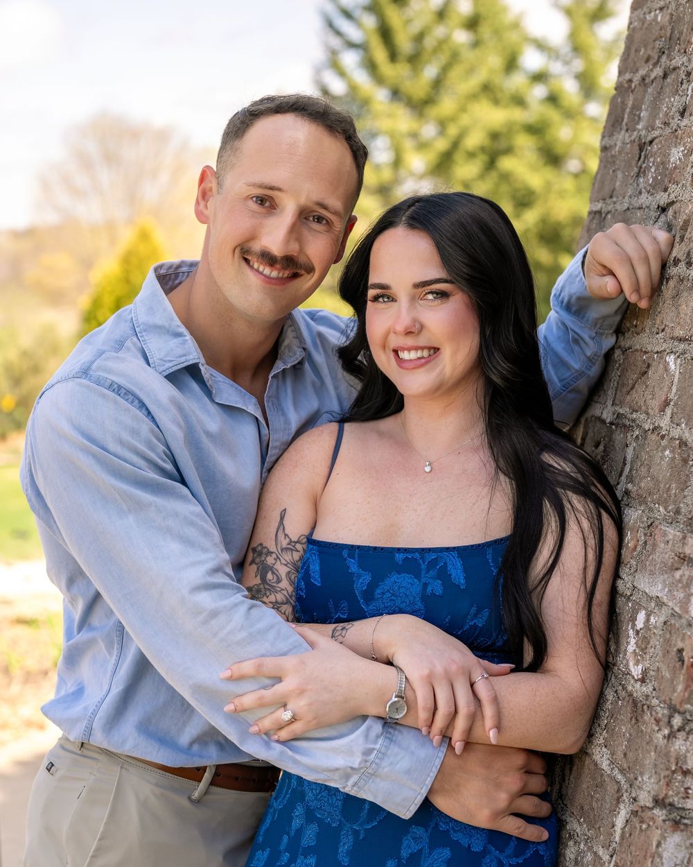 Happy engaged couple leaning against tree outdoors, woman in blue dress with tattoo, sunny day.