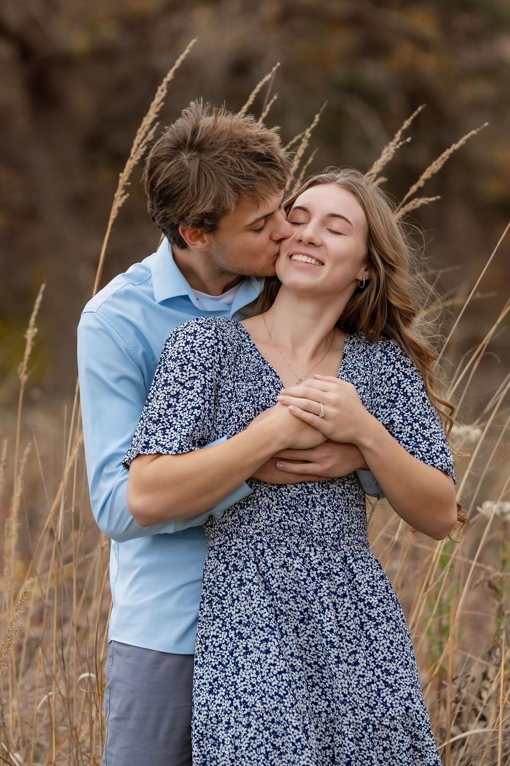 A smiling young woman in a blue floral dress is embraced and kissed on the cheek by a man in a blue shirt in a tall grass field.