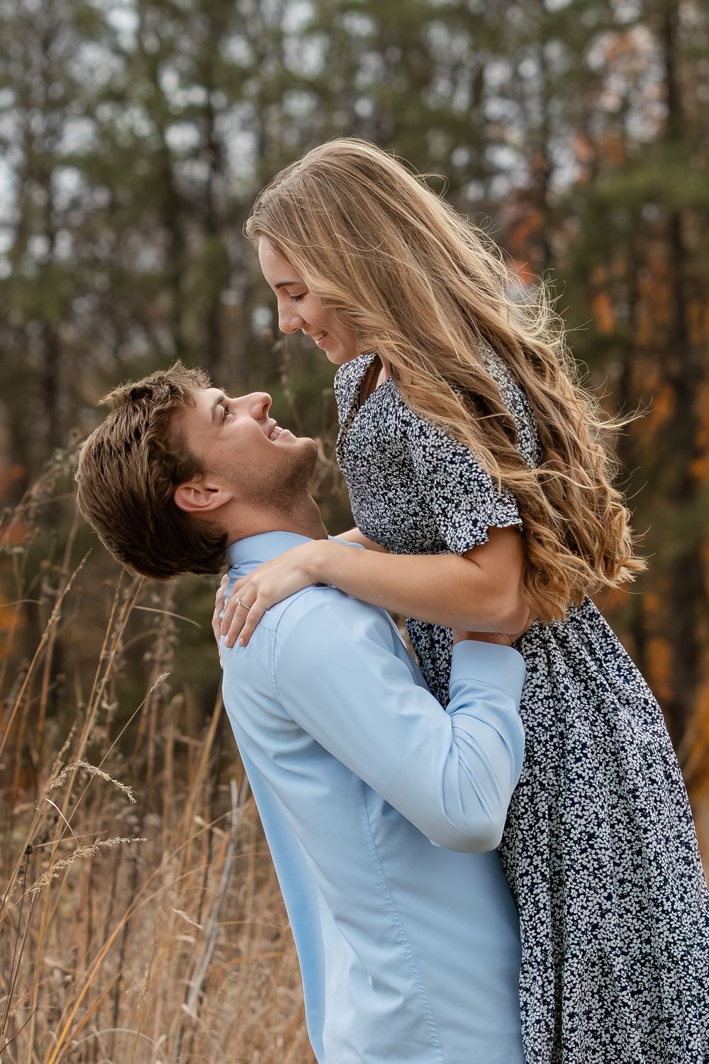 Smiling couple embracing in an autumn field as a man in a blue shirt lifts a woman in a floral dress with long wavy hair.