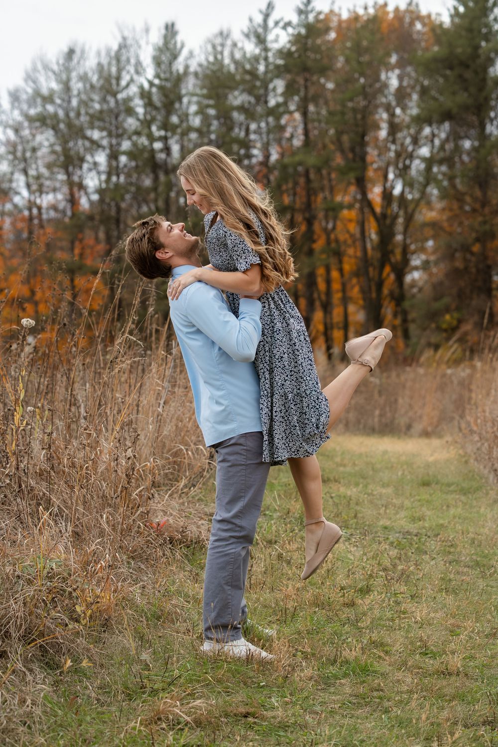 Man in a blue shirt lifts a smiling woman in a floral dress in a grassy field with colorful autumn trees behind them.
