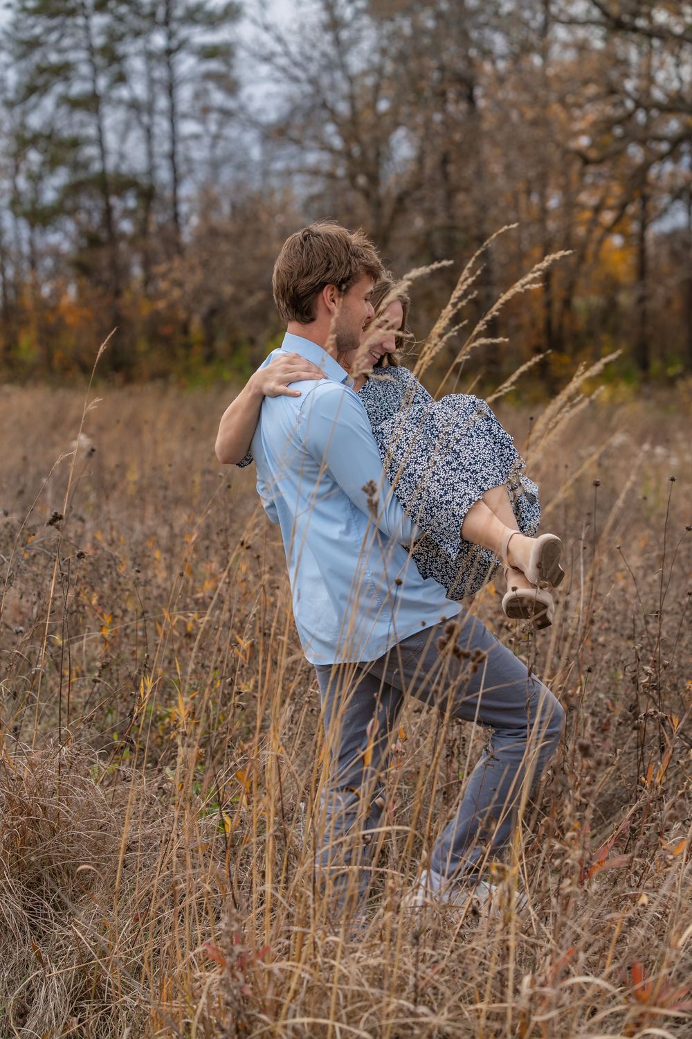 A young man in a blue shirt lifts and carries a woman in a floral dress through tall golden grass in an autumn field.