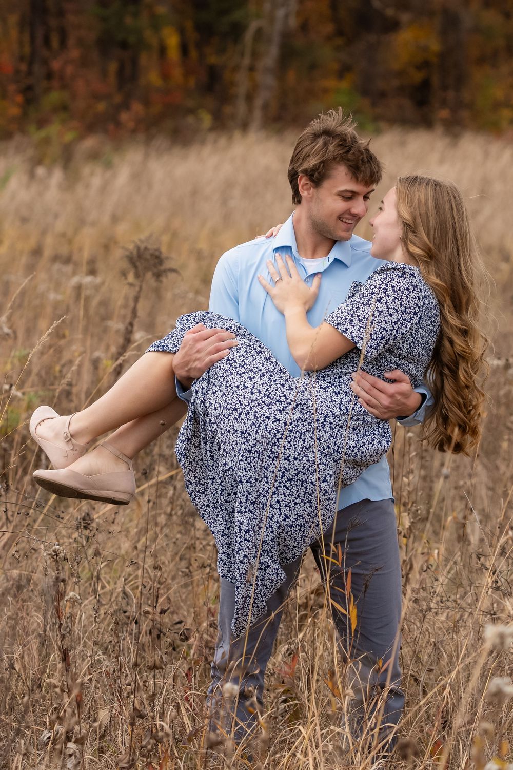 Smiling man in a blue shirt carries a woman in a floral dress through a tall autumn grass field during an engagement photo session.