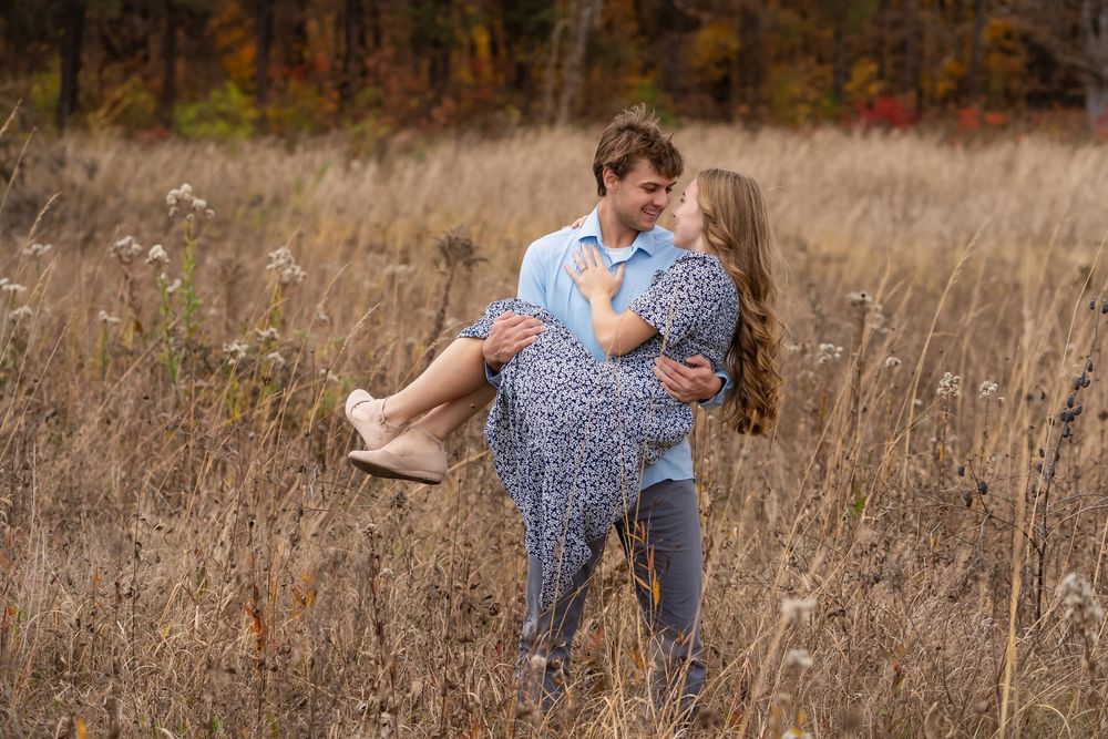 Man in a light blue shirt carries a smiling woman in a floral dress through a tall golden grass field with autumn trees behind.