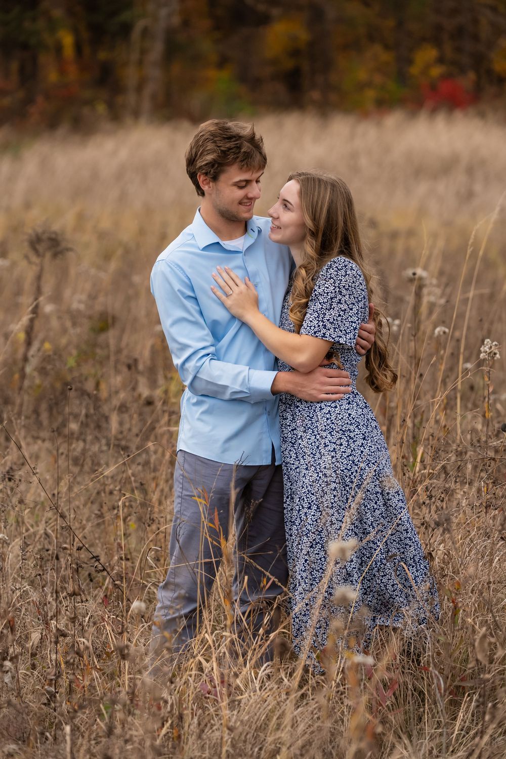 Smiling couple embraces in a tall golden grass field with autumn foliage behind them, a man in a blue shirt and a woman in a floral dress.